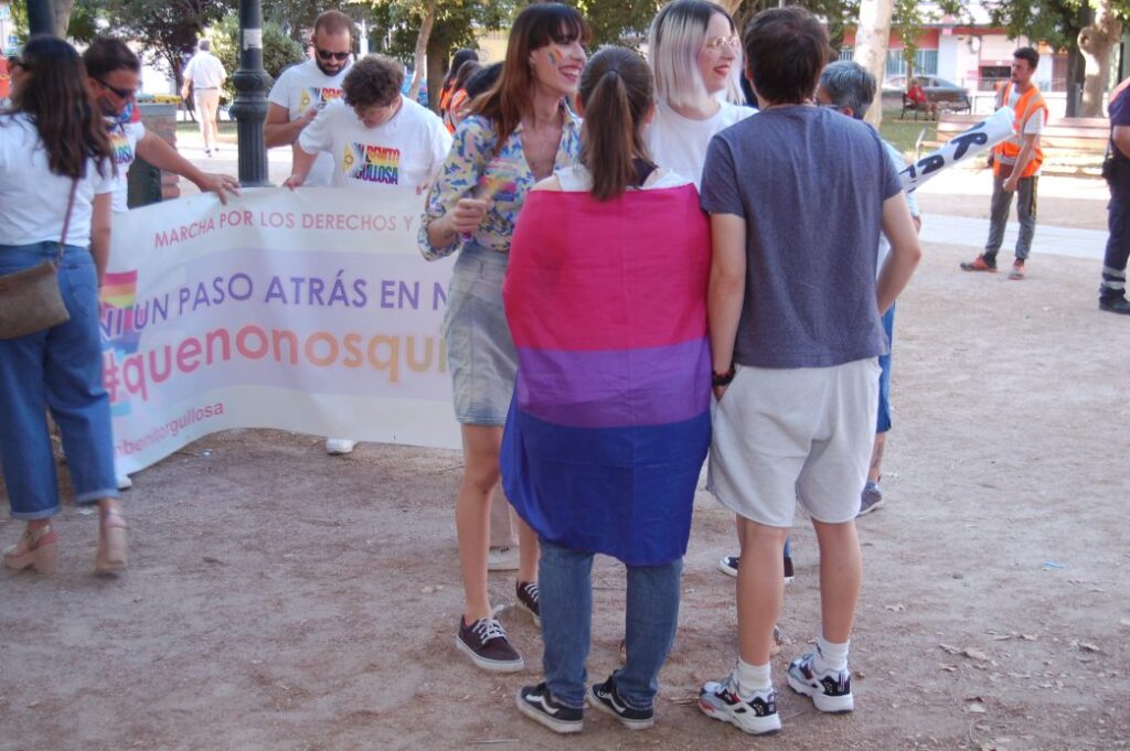 En un primer plano se puede ver un grupo de personas conversando en el que destaca una chica envuelta en una bandera bisexual. Al fondo está el colectivo "Don Benito Orgullosa" desplegando una pancarta.